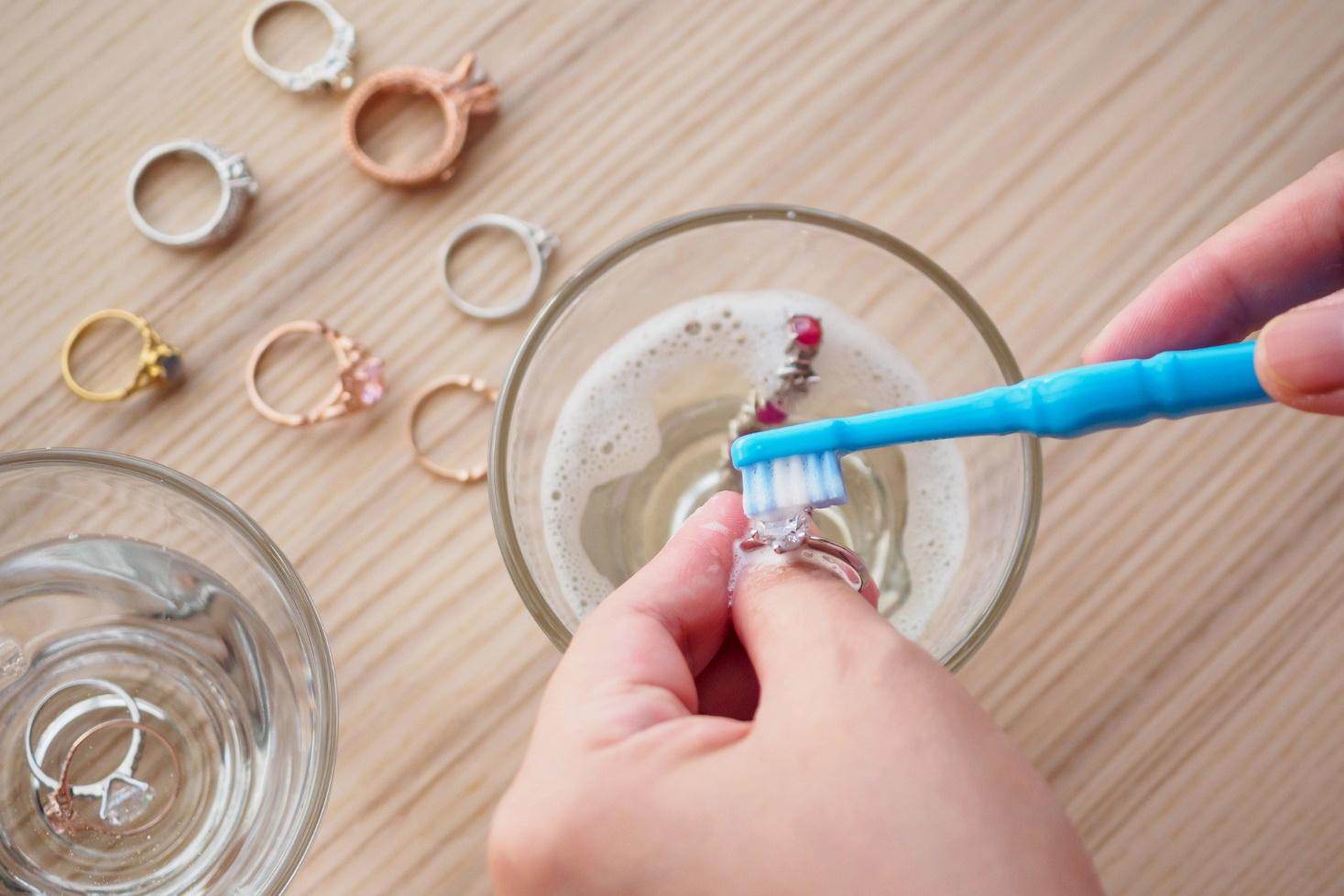 A person is cleaning jewelry using a toothbrush in soapy water.